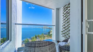 A balcony with wicker furniture, a view of the sea, and a cityscape in the background. - Amazing Studio with Oceanview - Pool - Beach - Gym (Puerto Vallarta)