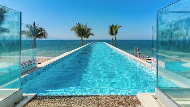 A clear blue pool with a view of the sea, palm trees, and a warning sign. - Amazing Studio with Oceanview - Pool - Beach - Gym (Puerto Vallarta)