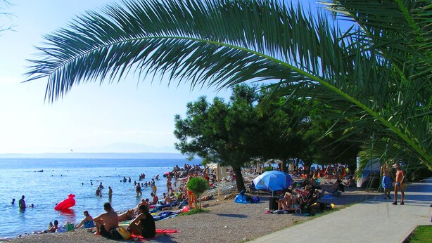 Ein Strand mit badenden und sonnenden Personen, einem klaren Himmel und ruhiger See.
