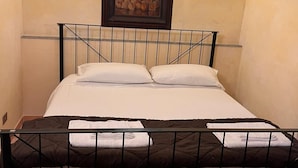 Un letto singolo con struttura in metallo nero, biancheria da letto bianca e trapunta marrone. Sopra il letto, un quadro incorniciato e un lampadario fissato alla parete.