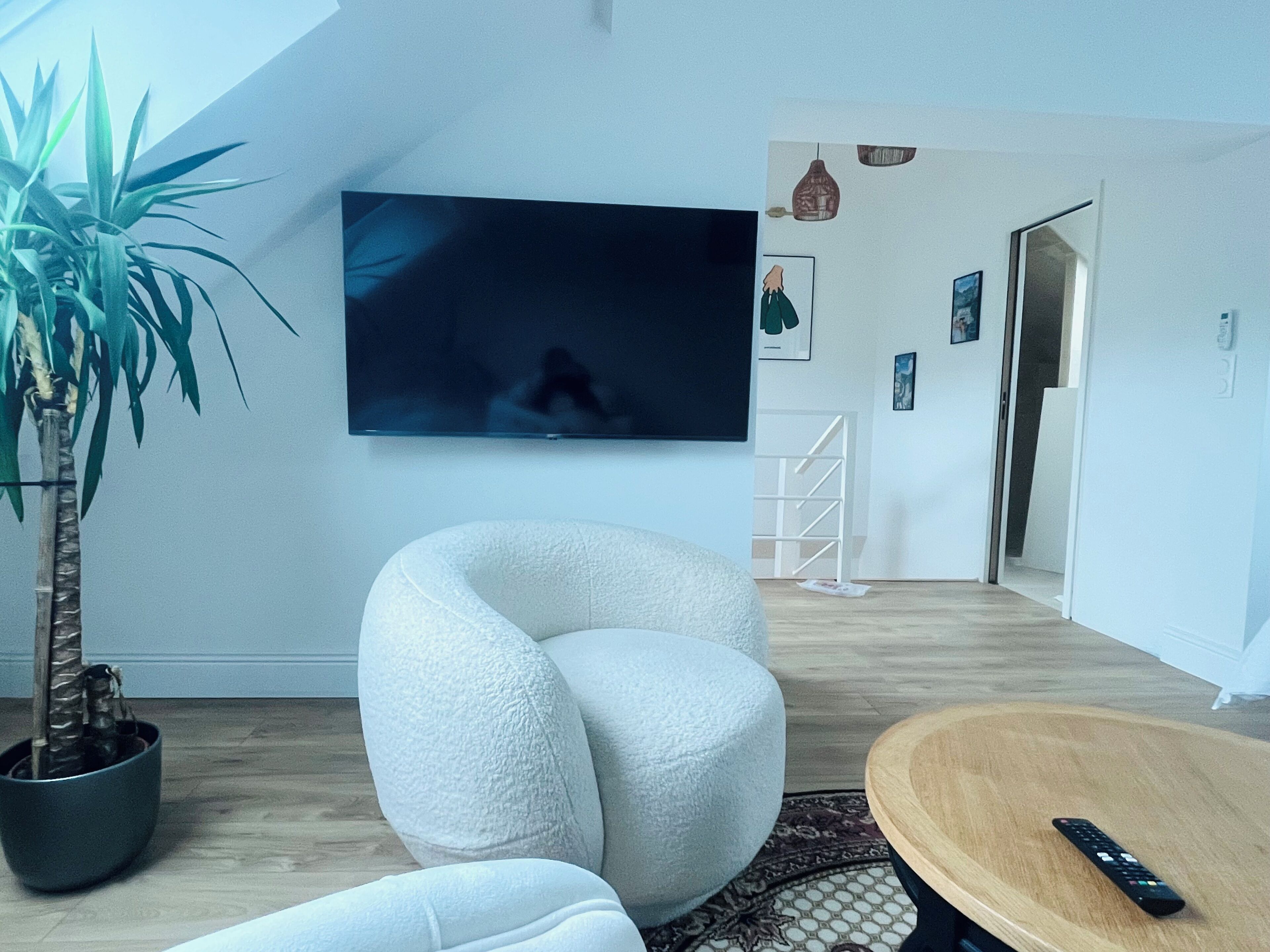 Un salon moderne avec un grand téléviseur à écran plat fixé au mur, un fauteuil blanc original, une table basse en bois et une plante en pot.
