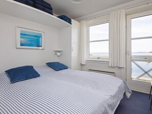Une chambre à coucher avec un lit, deux oreillers, une table de chevet, un tableau encadré, une fenêtre avec des rideaux et une vue sur la mer.