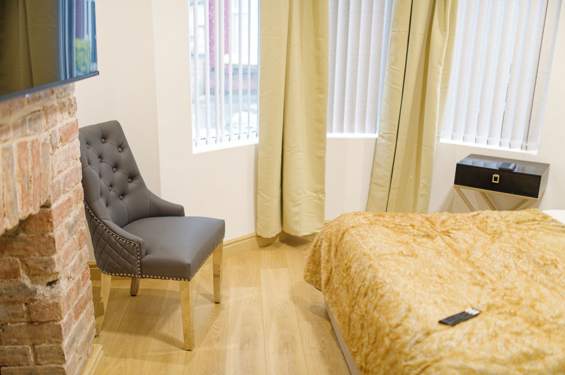 A bedroom with a tufted armchair, a bed with a textured blanket, a small bedside table, and a brick wall.