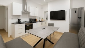 A modern kitchen with white cabinets, a stainless steel range hood, a microwave, and a built-in oven. There is a grey dining table with a remote control on it, and a grey sofa in the living area. - 4-BR Liverpool House Free Parking (Liverpool)