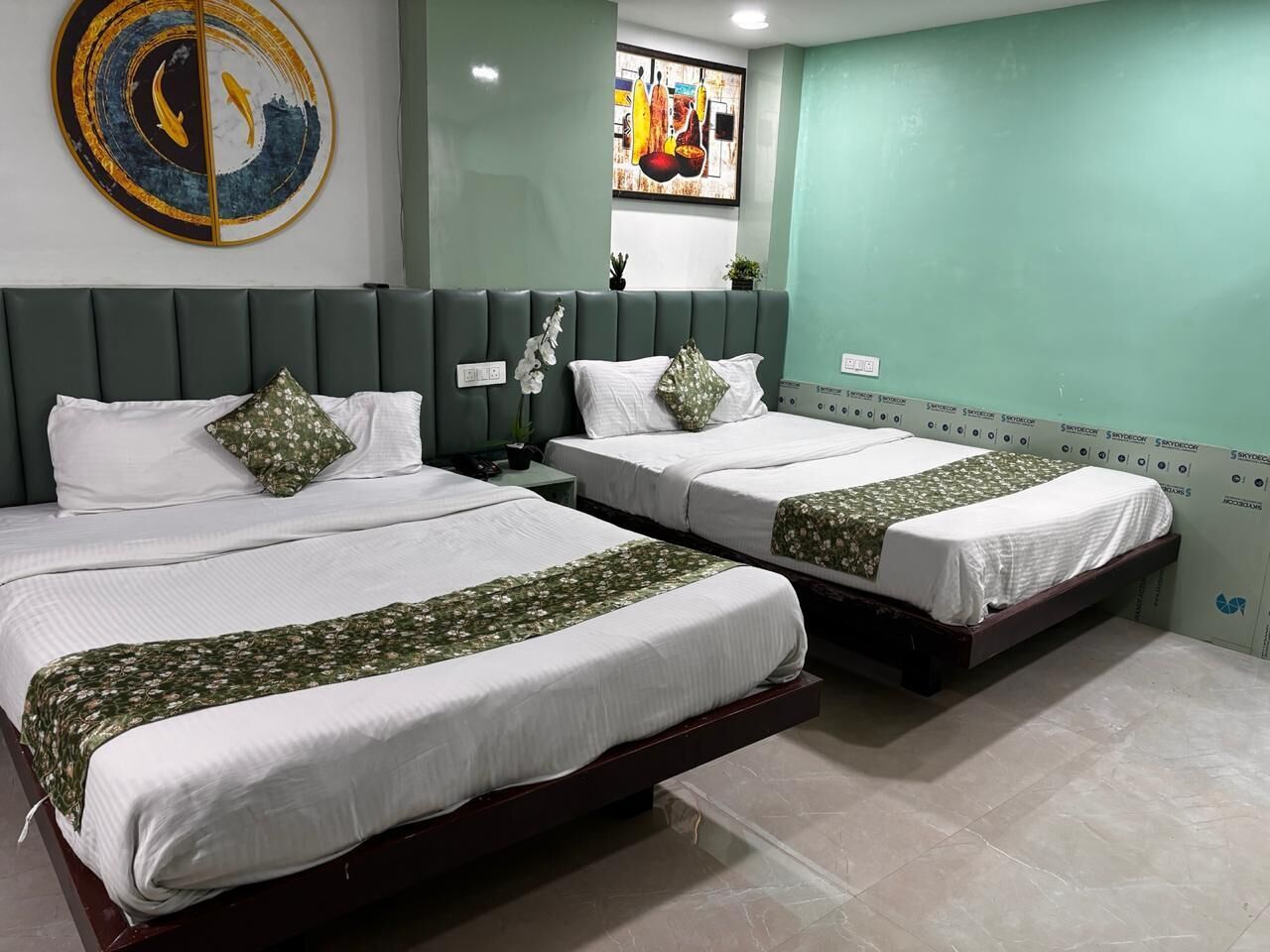 A hotel room with two beds, each with a green patterned bedspread and white pillows. There is a large abstract painting on the wall above the beds and a smaller painting below it.