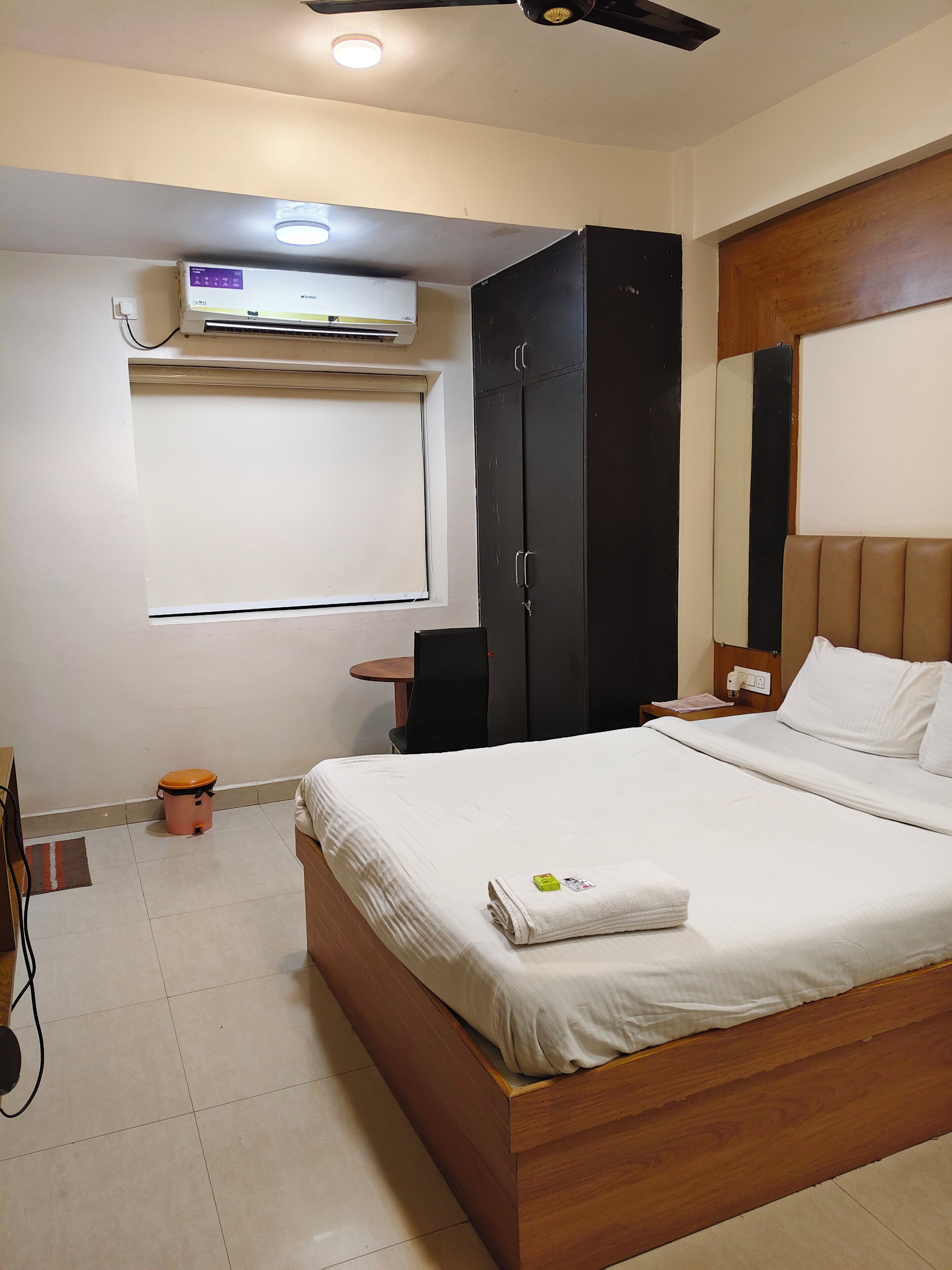 A hotel room with a bed, a wardrobe, a desk, a chair, and an air conditioning unit.