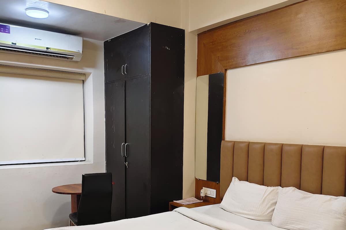Una cama bien hecha con cabecera de madera, una mesita de noche pequeña con una lámpara, un armario negro y un equipo de aire acondicionado en la pared.