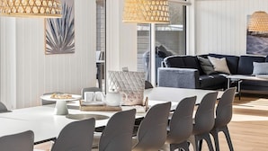 Uma área de jantar moderna com uma mesa branca, cadeiras cinzentas e um sofá preto ao fundo.