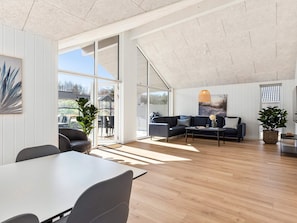 A spacious living area with a large window, a dark-colored sofa set, a dining table with chairs, and a wooden floor.