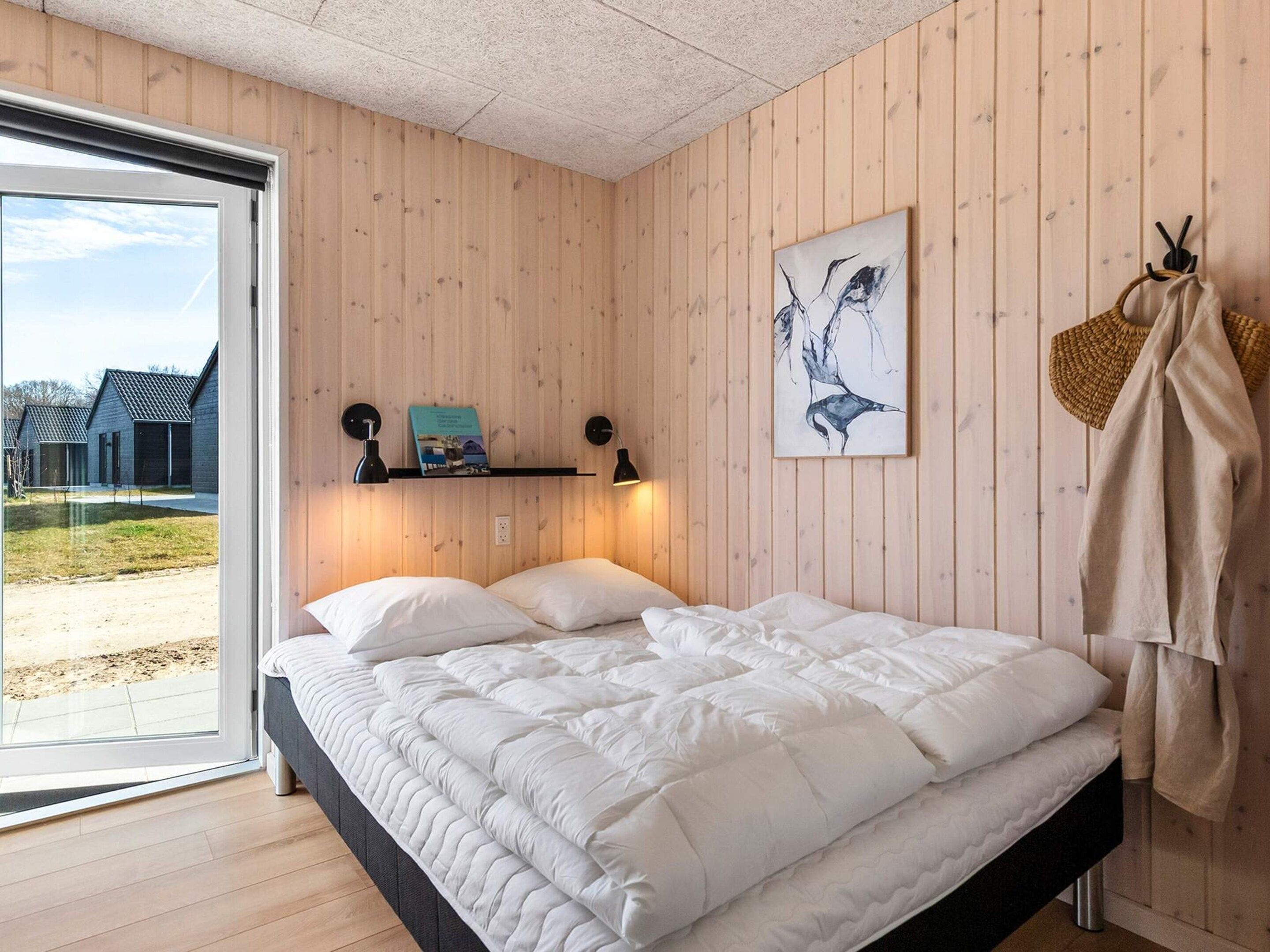 Quarto com cama, janela, estante de livros, uma obra de arte na parede e um cabideiro.