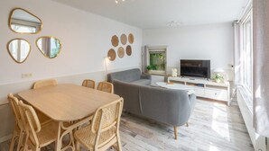 A modern living room with a grey sofa, a wooden dining table with wicker chairs, a flat-screen TV, and decorative wall art. - Spacious Duinland 188 Home With Veranda (Sint Maartensvlotbrug)