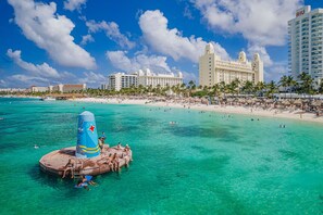 A beach scene with clear turquoise waters, a large inflatable structure, and a resort with multiple buildings in the background. - Grand Palm Luxury Villa w /Pool & Hot Tub by Bocobay (Noord)