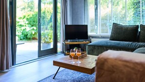 A modern living room with a sofa, a coffee table with drinks, a TV, and large windows with blinds. - Cozy forest cottage - cozy wooden cottage in the middle of nature on the Veluwe (Putten)