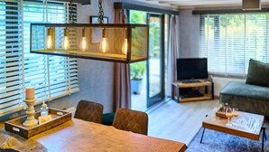 A modern living room with a wooden dining table, a hanging light fixture, a television, and a sofa. - Cozy forest cottage - cozy wooden cottage in the middle of nature on the Veluwe (Putten)