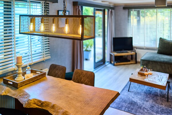 A modern living room with a wooden dining table, a hanging light fixture, a television, and a sofa.