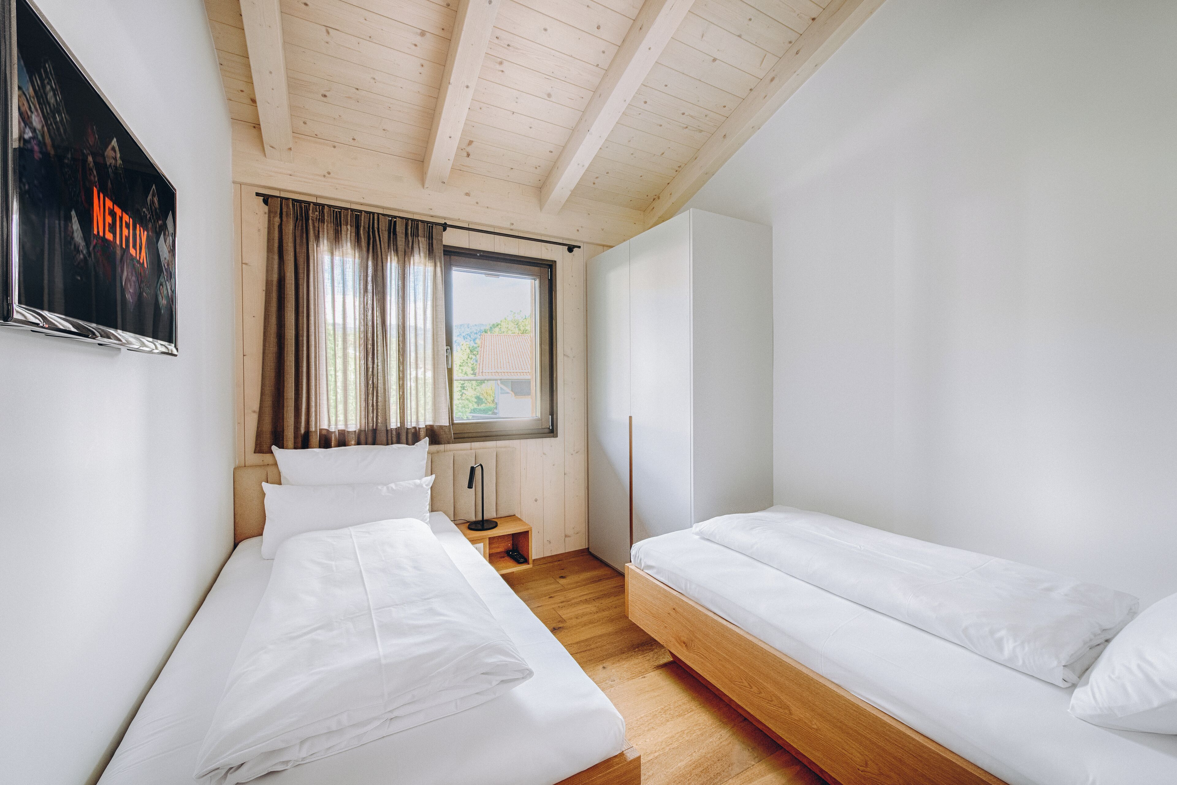 Ein Schlafzimmer mit zwei Einzelbetten, einer Holzbalkendecke, einem Fenster mit Vorhängen und einem an der Wand befestigten Fernseher, der 'NETFLIX' anzeigt.