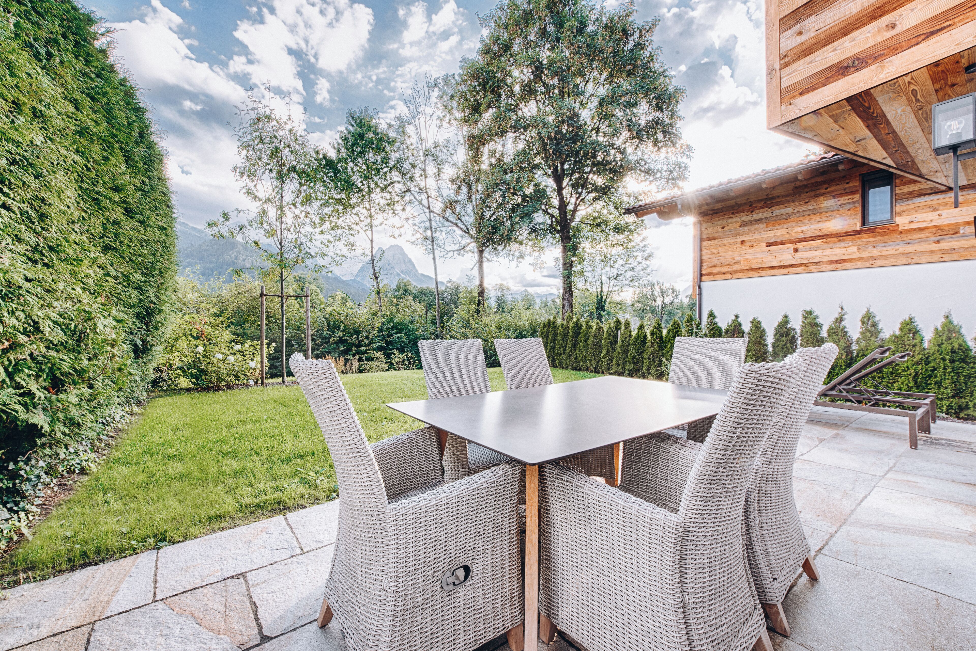 Une terrasse extérieure avec une table rectangulaire et des chaises en osier, entourée d’un jardin luxuriant et offrant une vue sur les montagnes.