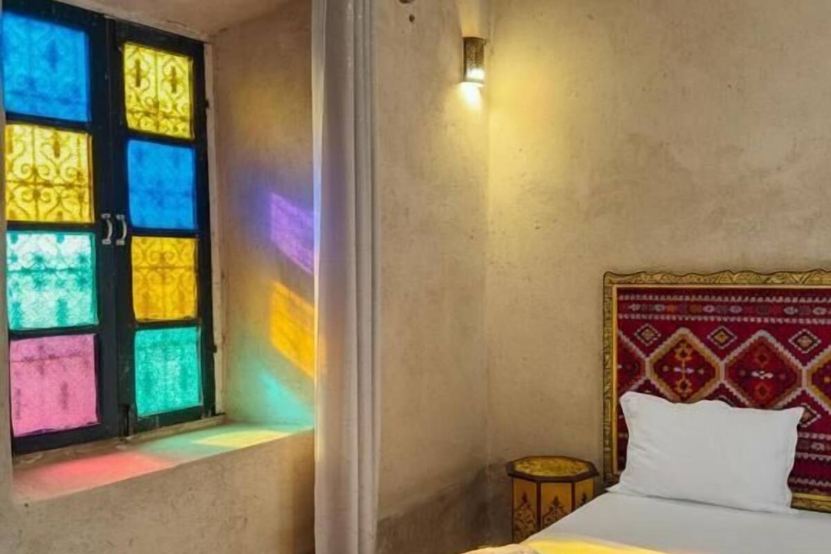 A bedroom with a bed, a window with colorful stained glass, and a wall-mounted light.