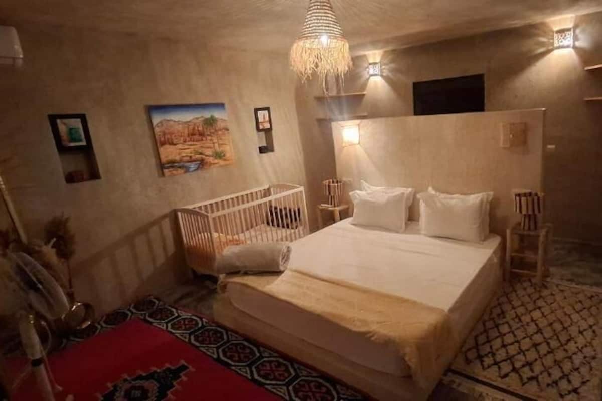 A bedroom with a large bed, a crib, a patterned rug, and wall-mounted lighting.