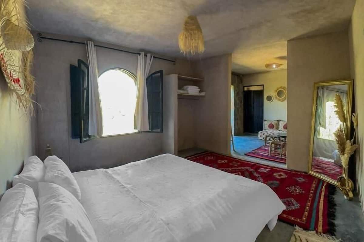 A bedroom with a large bed, a window with curtains, a red carpet, and a mirror.