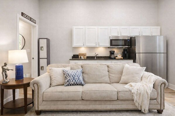 Ein Wohnzimmer mit einem beigen Sofa, einem kleinen Tisch mit Lampe, einer Küchenzeile mit weißen Schränken, einer Mikrowelle und einem Kühlschrank.