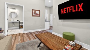 A modern living room with a wooden coffee table, a green ottoman, a flat-screen TV with 'NETFLIX' on it, a framed picture on the wall, and a kitchen area in the background. - Legends Retreat: A spacious stay for large groups (Louisville)