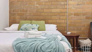 A neatly made bed with a green blanket, a pillow with a floral pattern, and a white pillow. There is a small wooden table beside the bed with a white basket underneath. - Legends Retreat: A spacious stay for large groups (Louisville)