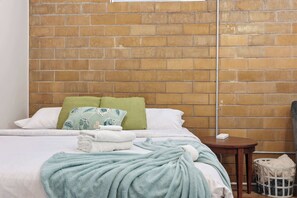 A neatly made bed with a green blanket, a pillow with a floral pattern, and a white pillow. There is a small wooden table beside the bed with a white basket underneath.