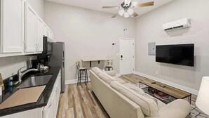 A modern kitchen with white cabinets, a black countertop, and a built-in oven. There is a wooden dining table with two chairs, a beige sofa, and a flat-screen TV mounted on the wall. - Legends Retreat: A spacious stay for large groups (Louisville)