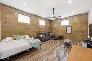 A modern room with a brick wall, a bed, a sofa, a ceiling fan, and a small table. - The Booker Noe Suite | Brick Home w/ Modern Design (Louisville)