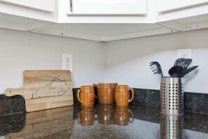 A kitchen counter with a wooden cutting board, three wooden mugs, and a utensil holder. - The Booker Noe Suite | Brick Home w/ Modern Design (Louisville)
