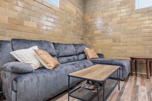 A corner room with a grey sofa, two cushions, a wooden coffee table, and a small wooden side table. - The Booker Noe Suite | Brick Home w/ Modern Design (Louisville)