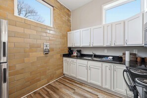 A kitchen with white cabinets, a brick wall, a stainless steel refrigerator, and a black countertop. - The Booker Noe Suite | Brick Home w/ Modern Design (Louisville)