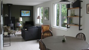A modern living room with a black sofa, a dining table, and a large window with a view of greenery. - La Bellevue forest-edge cottage (Hénouville)