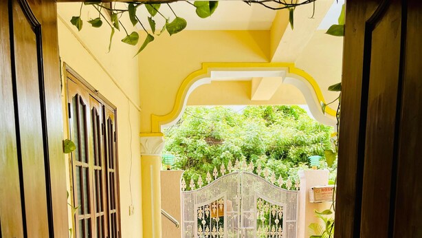 Une porte en bois ouverte donne sur une cour dallée, agrémentée d’une grille décorative, de plantes en pot et d’un mur jaune.