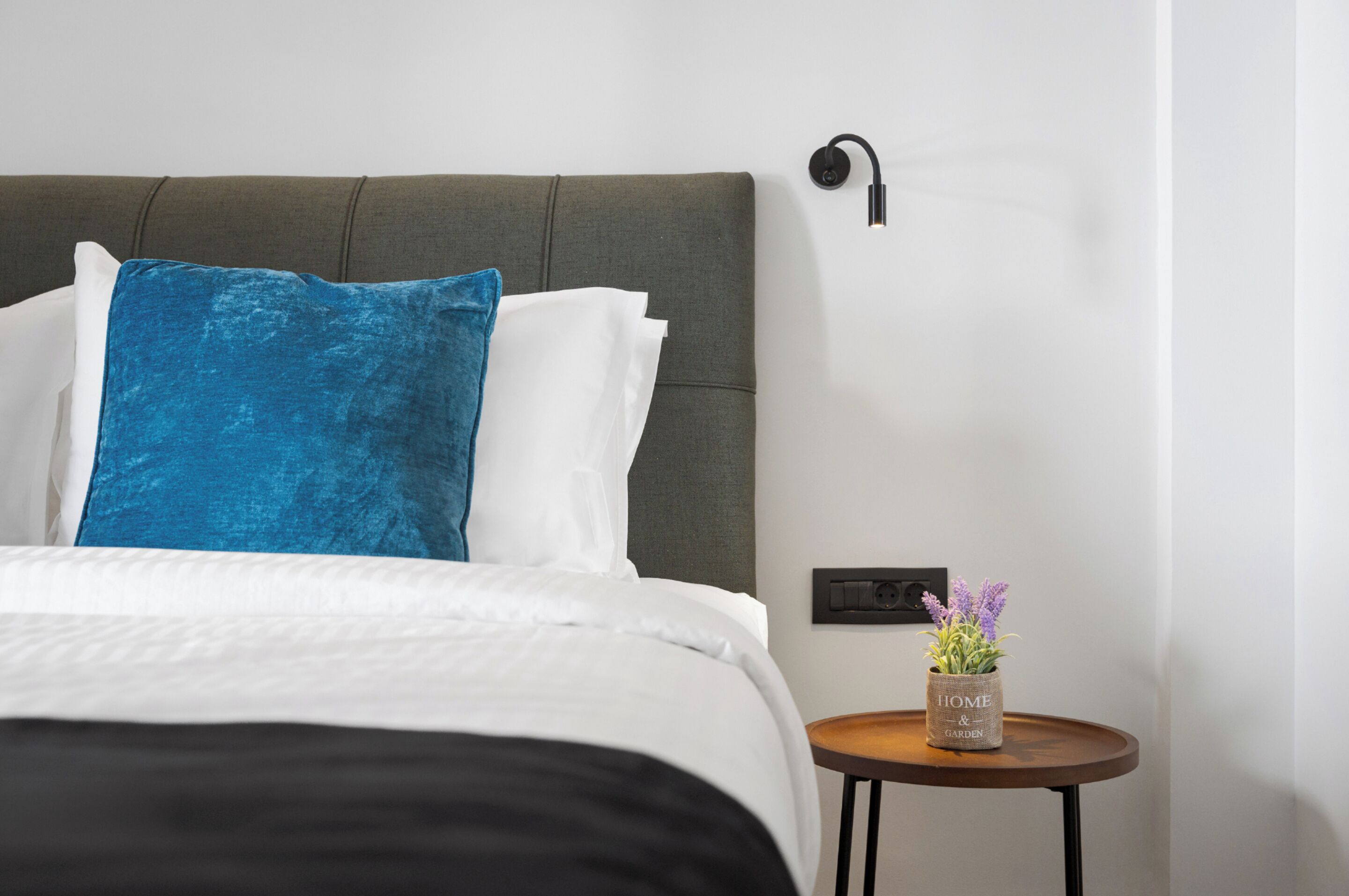 A neatly made bed with white and blue pillows, a grey upholstered headboard, a small round table with a potted plant, and a wall-mounted light fixture.