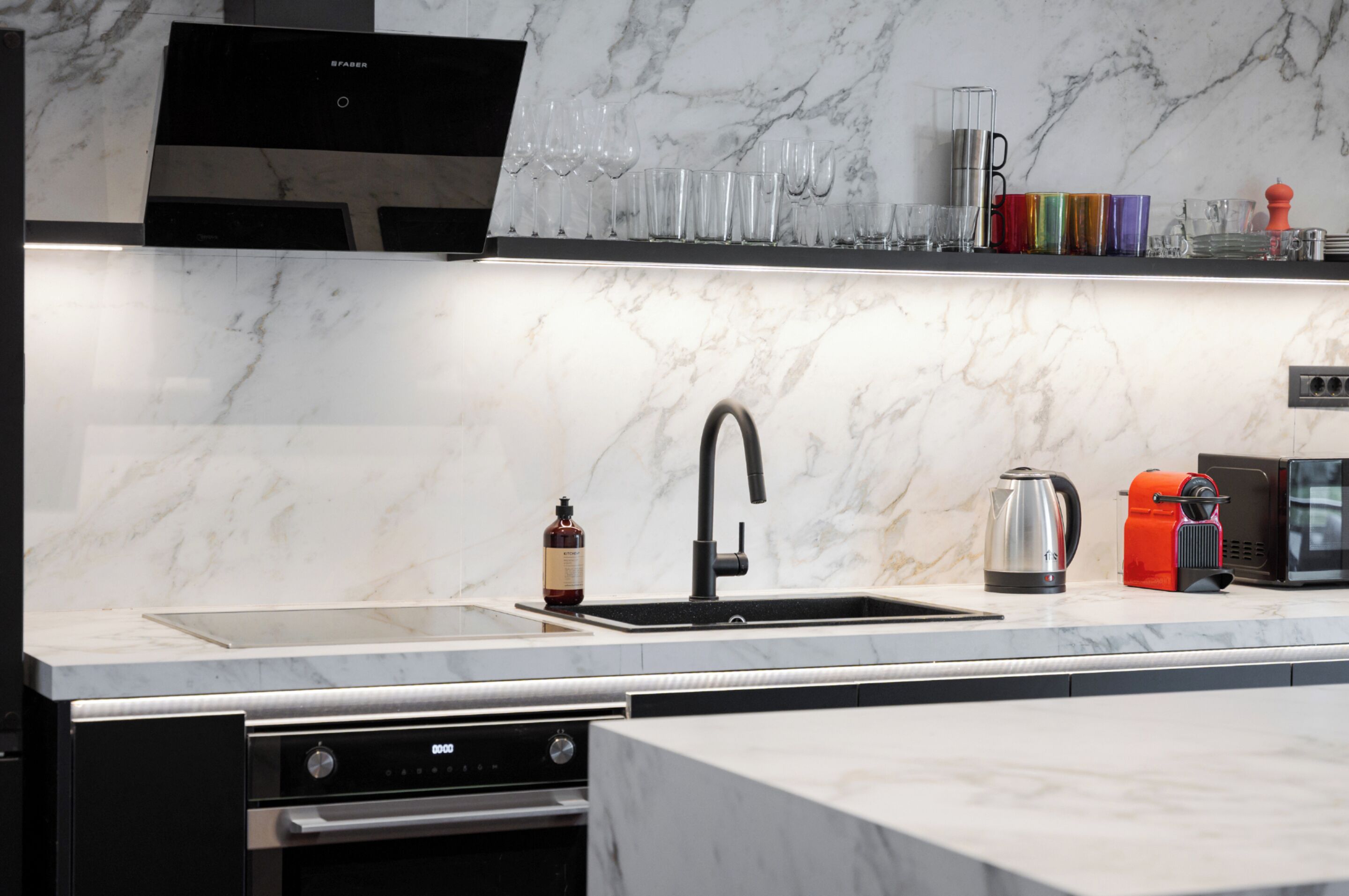 A modern kitchen with a marble countertop, stainless steel appliances, and a variety of glassware and kitchen tools.