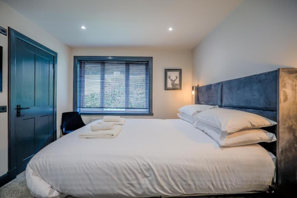 A neatly made bed with white linens, a dark headboard, and a window with blinds.