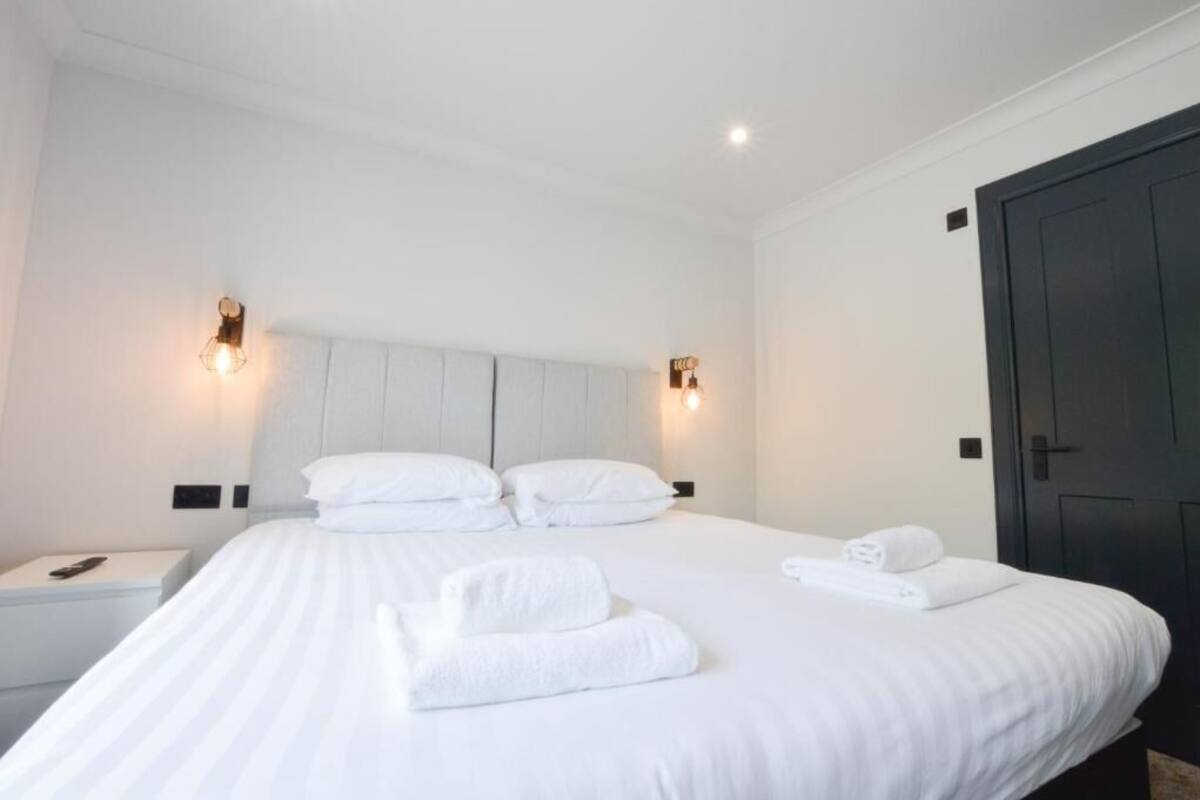 A neatly made bed with white linens and towels, a bedside table with a lamp, and a dark door.