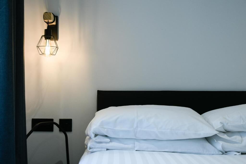 Una cama bien hecha con sábanas blancas y cabecera negra, una lámpara fijada a la pared y un enchufe en la pared.