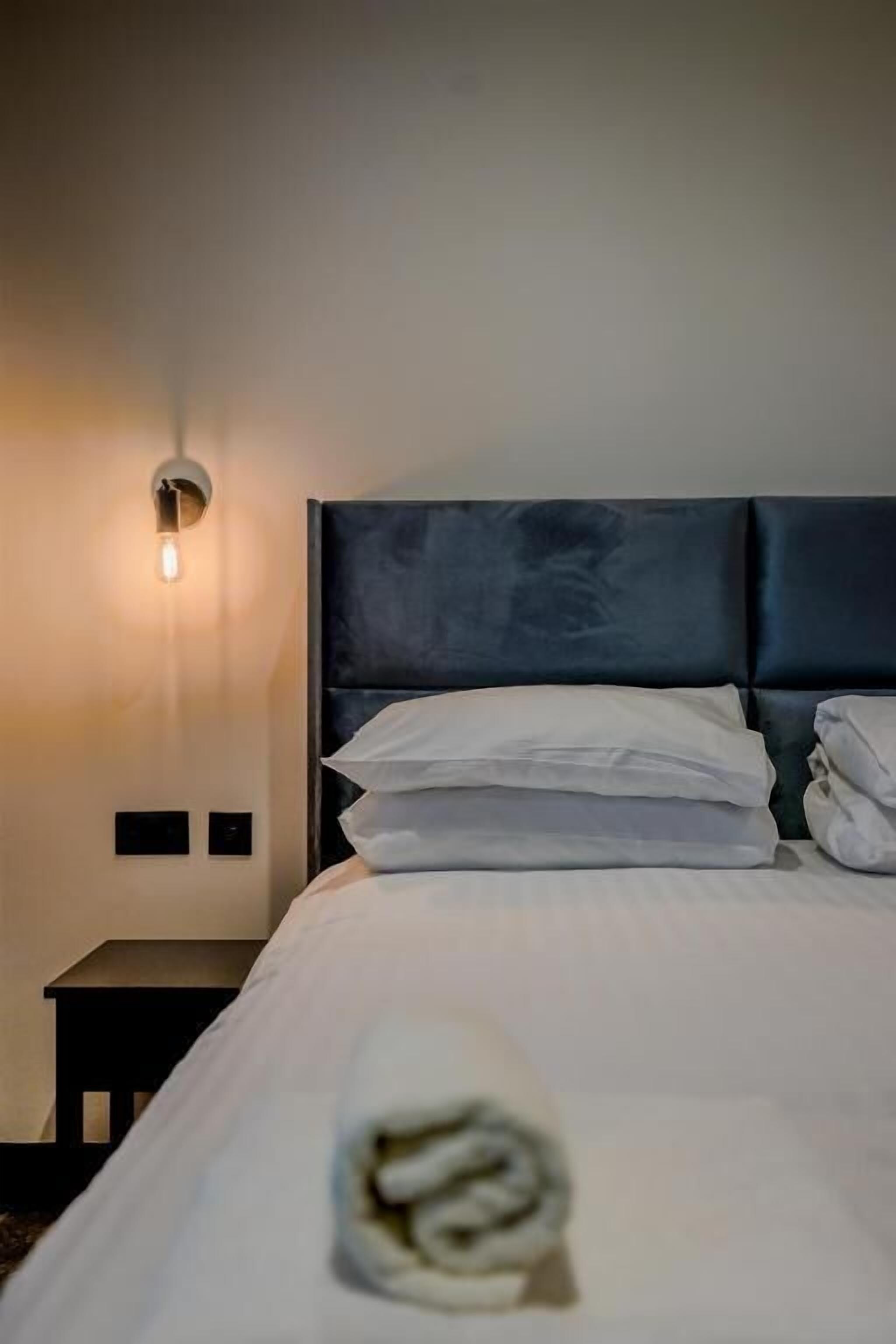 A neatly made bed with white linens and a rolled towel, a dark upholstered headboard, and a wall-mounted light fixture.