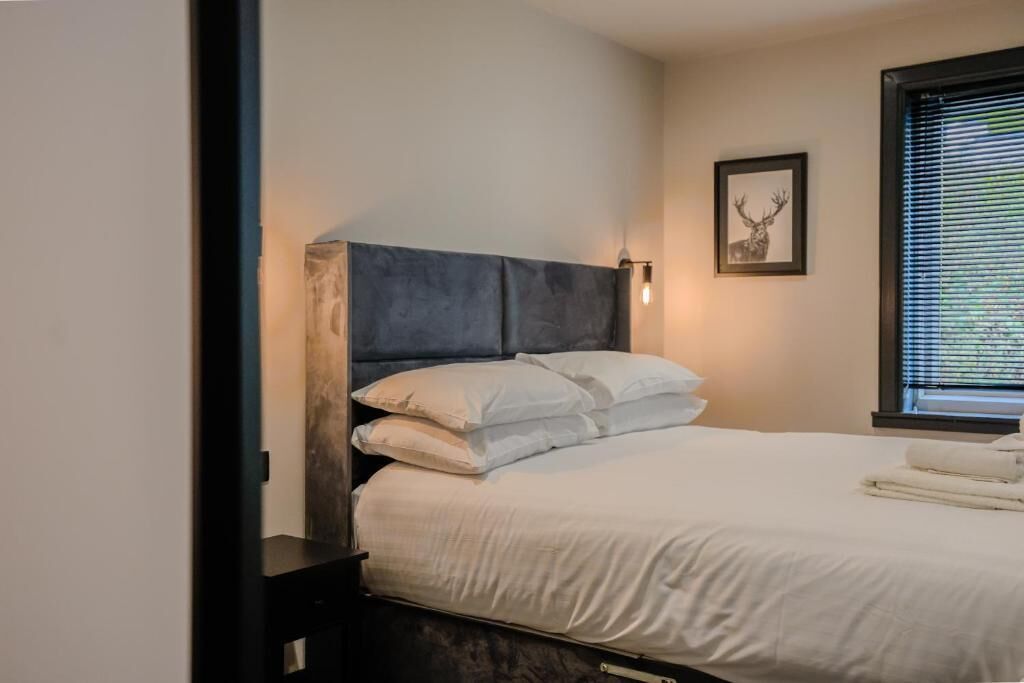 A neatly made bed with white linens and a dark headboard, a framed picture on the wall, and a window with blinds.
