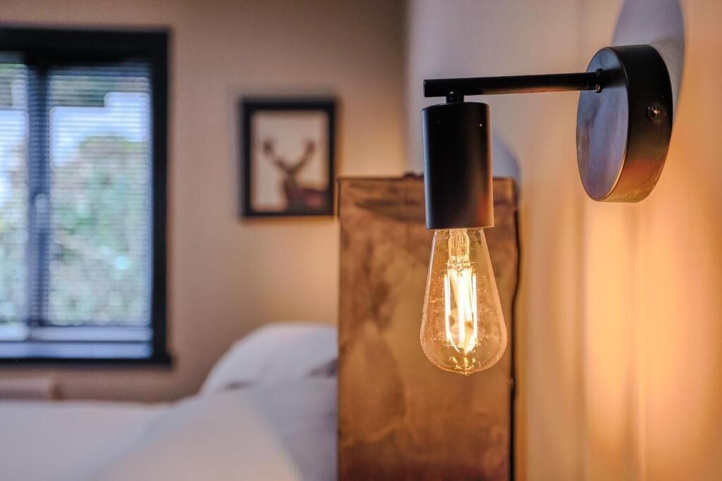 A room with a bed, a window with blinds, a framed picture on the wall, and a wall-mounted light fixture with a visible filament bulb.