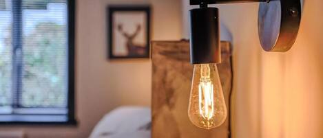 A room with a bed, a window with blinds, a framed picture on the wall, and a wall-mounted light fixture with a visible filament bulb.