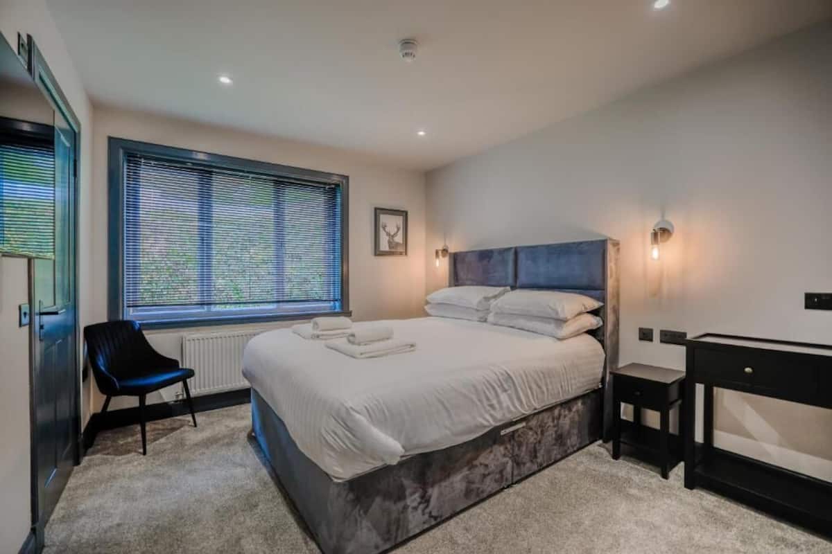 A modern bedroom with a large bed, a chair, a desk, and a window with blinds.