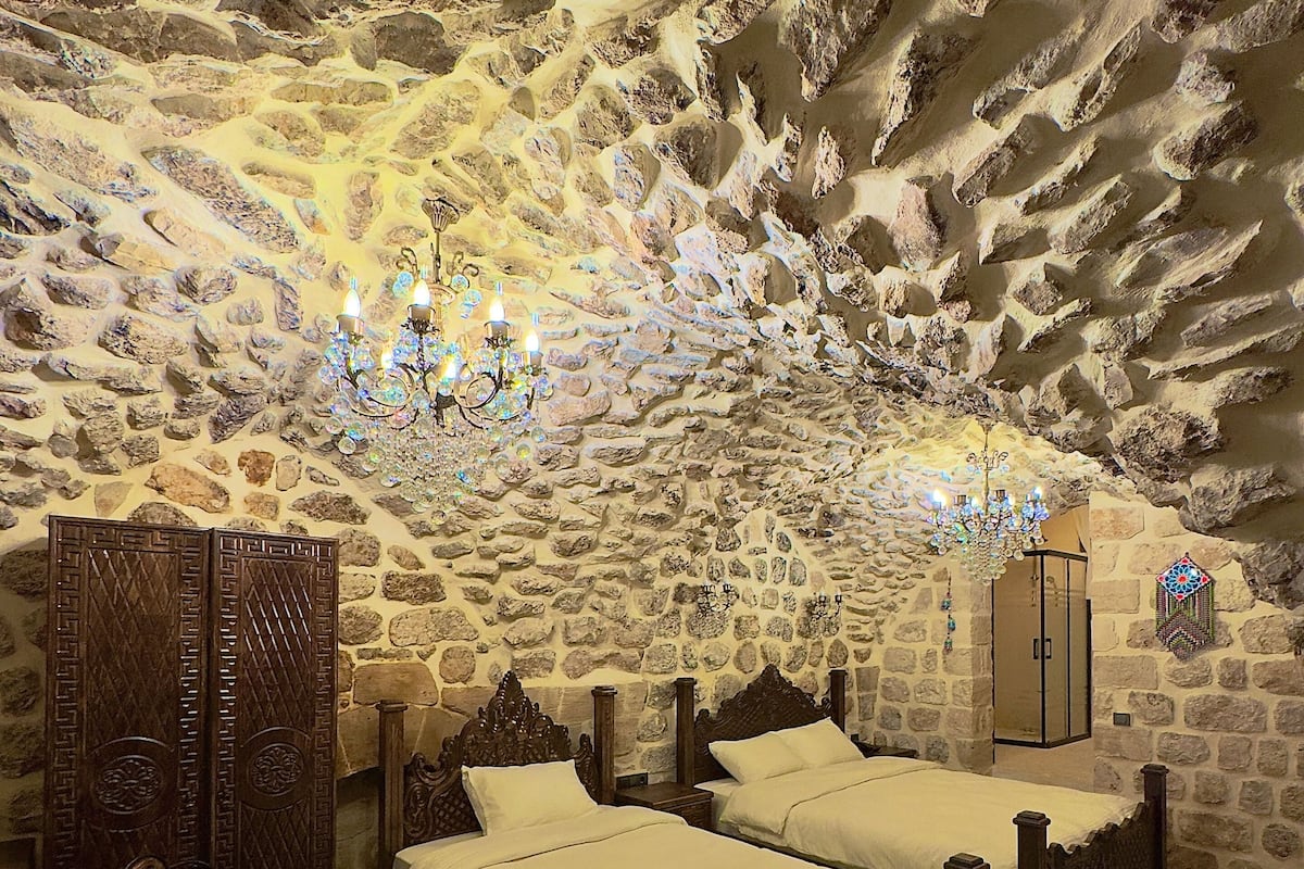 A room with stone walls and a high ceiling, featuring two wooden beds, a small round table, and a patterned rug.