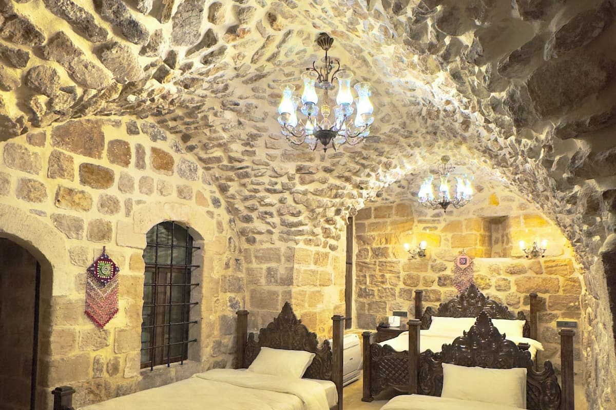 A stone-walled room with two beds, a chandelier, and a round wooden table.