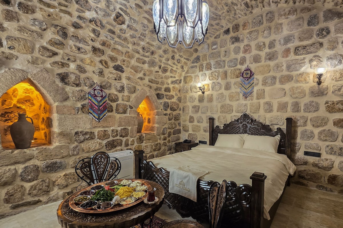 A stone-walled room with a bed, a table with a traditional tray, and decorative wall hangings.