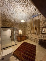 A stone-walled bathroom with a glass shower enclosure, a copper sink, and a patterned rug.