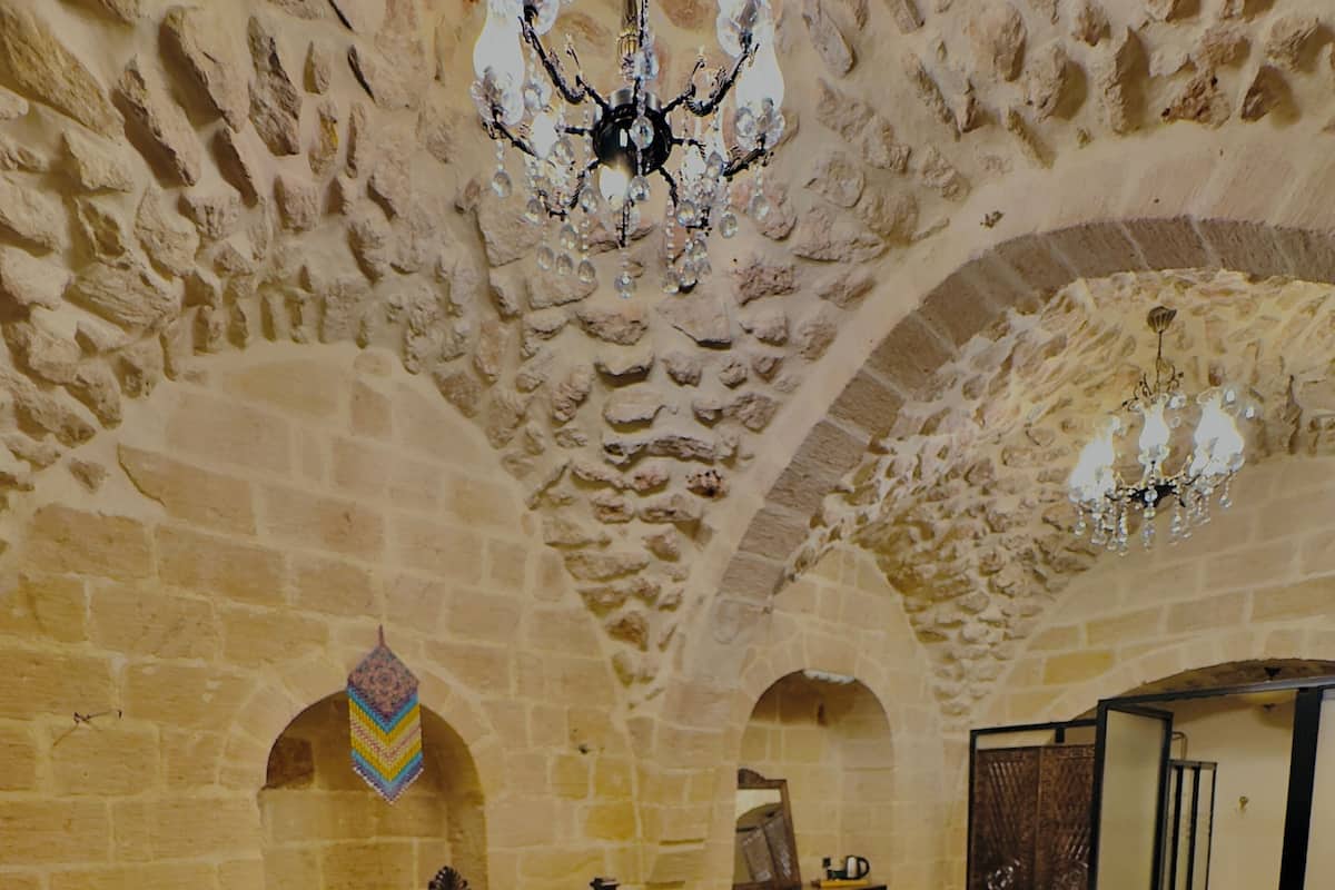A room with a large bed, wooden headboard, and a chandelier. There are arched stone walls and a small table with chairs.
